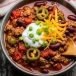 A bowl of hearty homemade chili filled with ground beef, beans, tomatoes, and bell peppers, topped with melted cheddar cheese, green onions, and tortilla chips — the ultimate comforting chili recipe for cozy nights.
